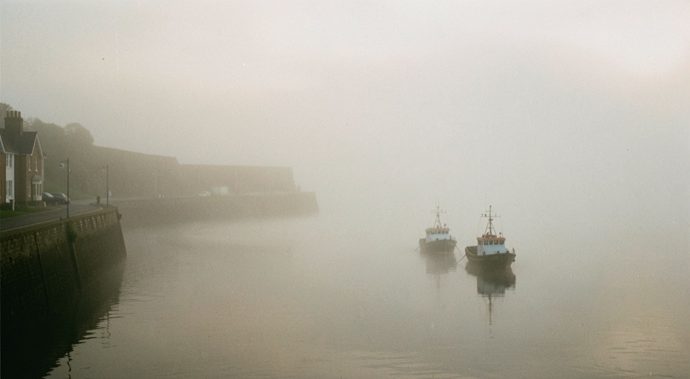 Sea fog filling a small Fife harbour at dawn, the far harbour wall dissolving into white, two moored boats in silhouette, soft flat light, wide framing