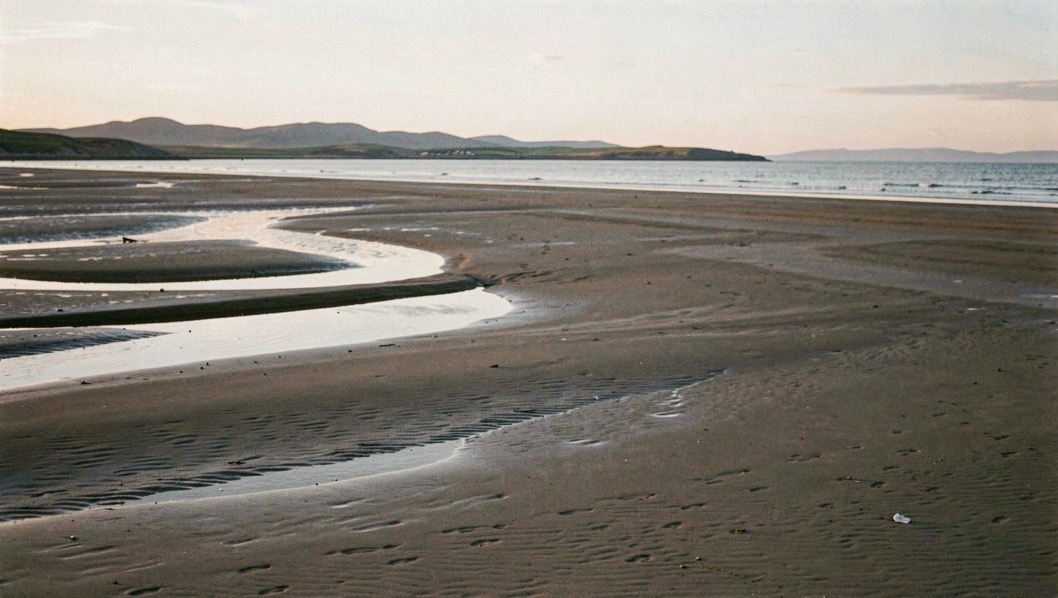 A wide plain of wet sand at low water on the Solway with shallow draining channels, the Galloway hills low on the far horizon, thin evening light along the ridges of sand, wide horizontal framing