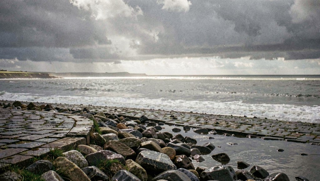 A wide shingle bank on the Suffolk coast just after a squall has passed, wet stones running from the bottom of the frame to a grey sea, a bright band of low sunlight along the waterline, dark cloud filling the sky above